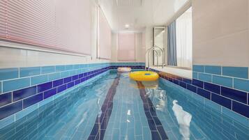 Una piscina interior con azulejos azules y blancos, dos aros inflables y un ventanal con cortinas.