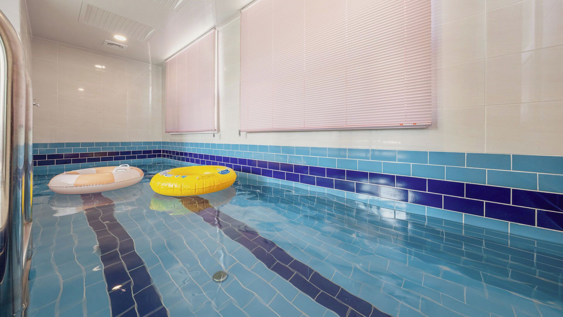 A swimming pool with blue and white tiles, two inflatable rings, and a window with blinds.