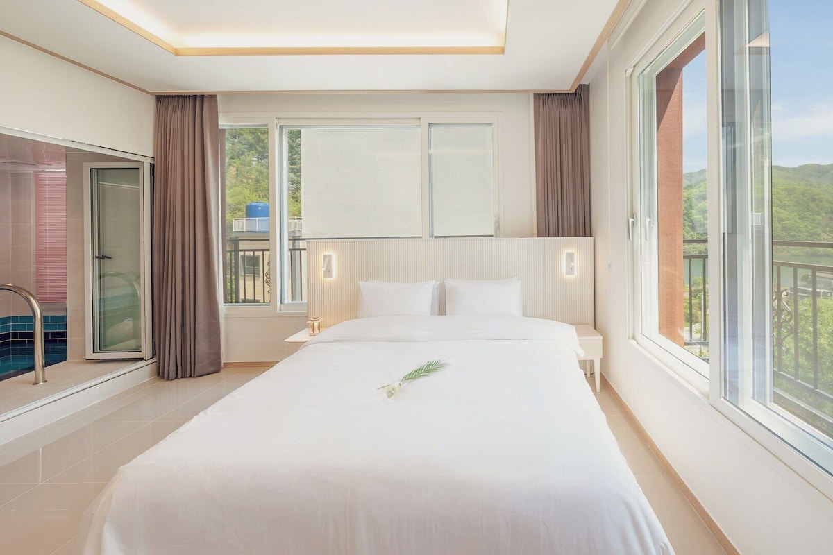 A hotel room with a large bed, a balcony with a view of greenery, and a sliding glass door leading to a pool area.