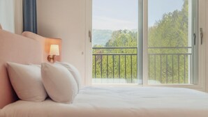 A bedroom with a bed, pillows, a nightstand with a lamp, and a balcony with a view of trees. - Gapyeong Laonjena Pool Villa (Gapyeong)
