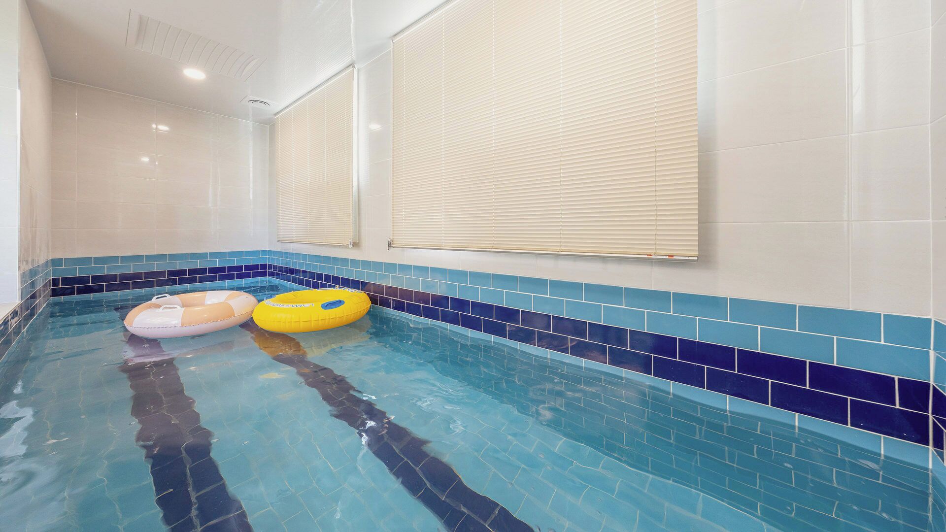 Piscina interna com paredes azulejadas em azul, dois flutuadores infláveis e teto com iluminação embutida.