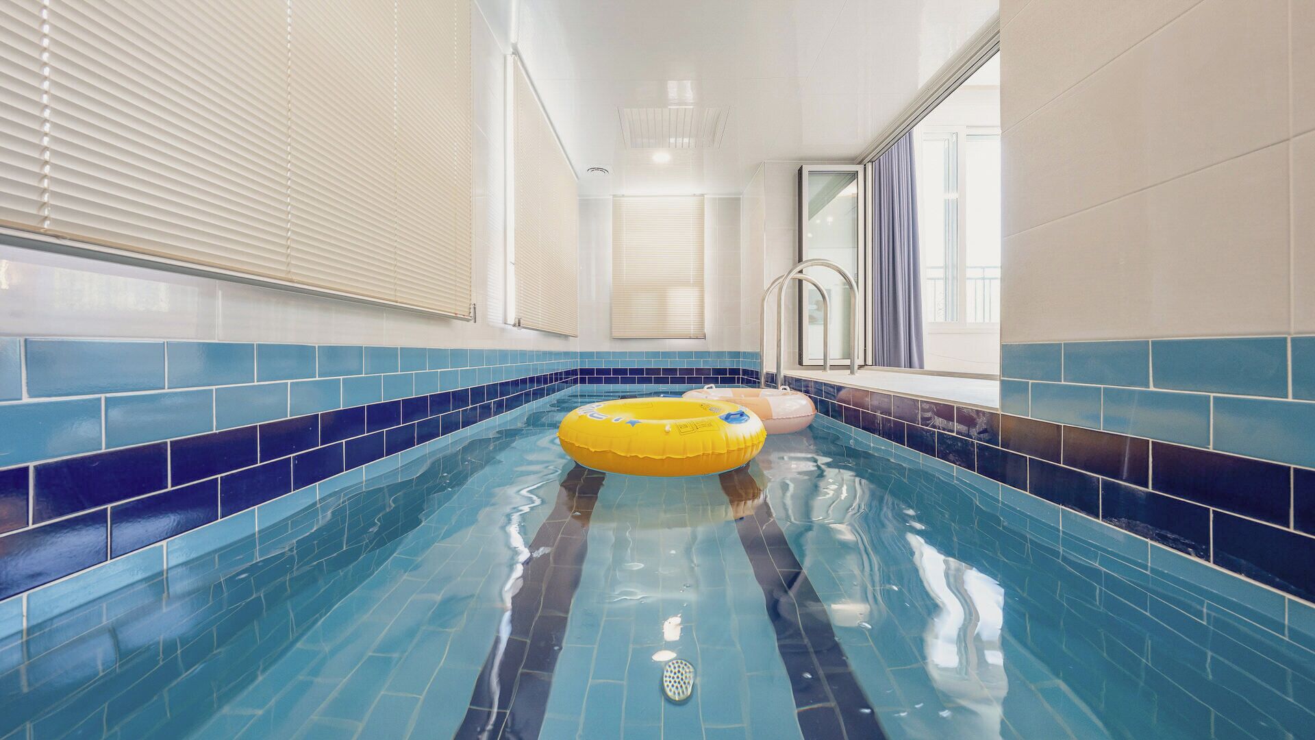 Ein Hallenschwimmbad mit blauen Fliesen, einem gelben Schwimmreifen und einem Fenster mit Vorhängen.