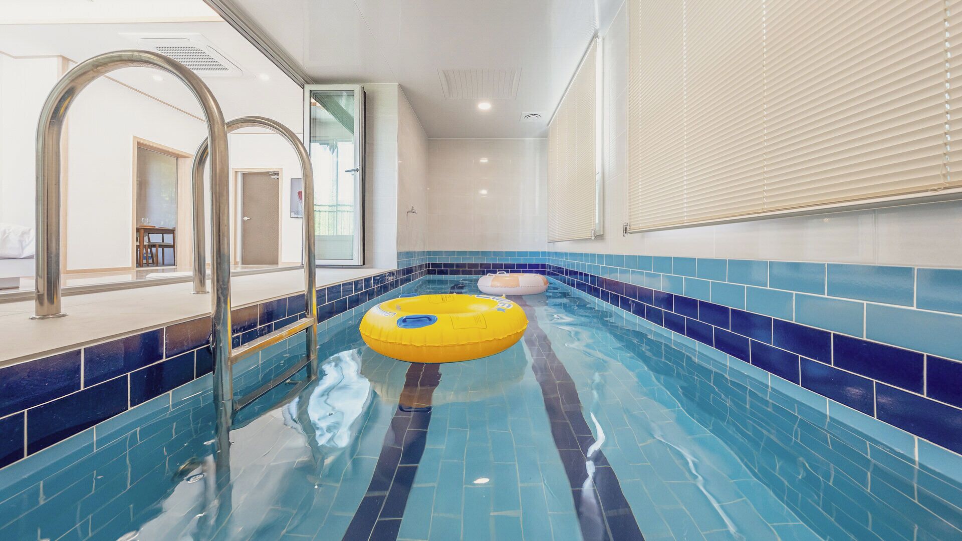 Une piscine intérieure aux carreaux bleu clair, un flotteur jaune et une porte vitrée donnant sur une pièce.