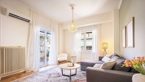 A modern living room with a sofa, armchair, and coffee table. There is a large window with curtains, a radiator, and a hanging light fixture.