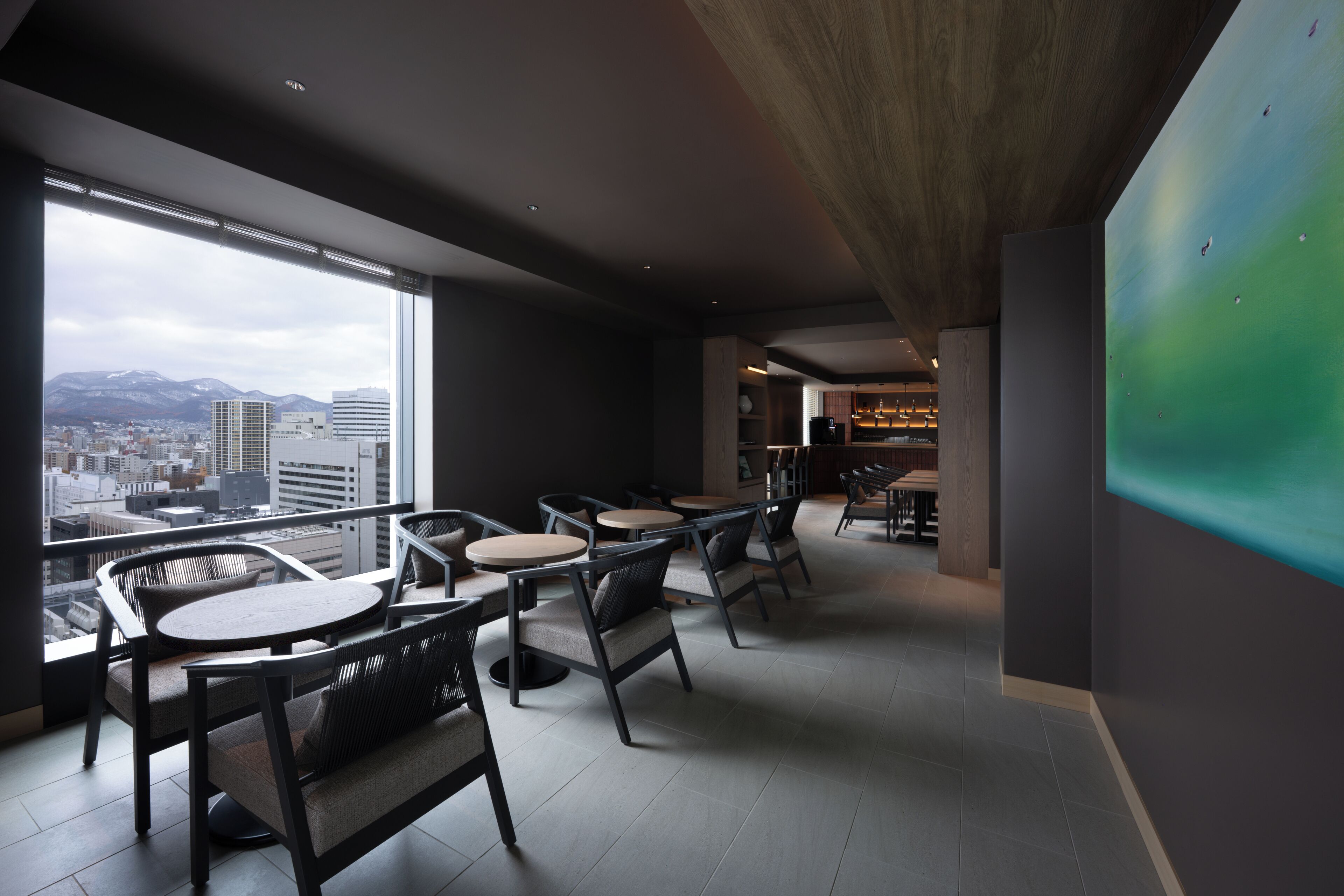 A modern interior with a city view, featuring a dining area with round tables and chairs, a bar area, and a large abstract painting on the wall.