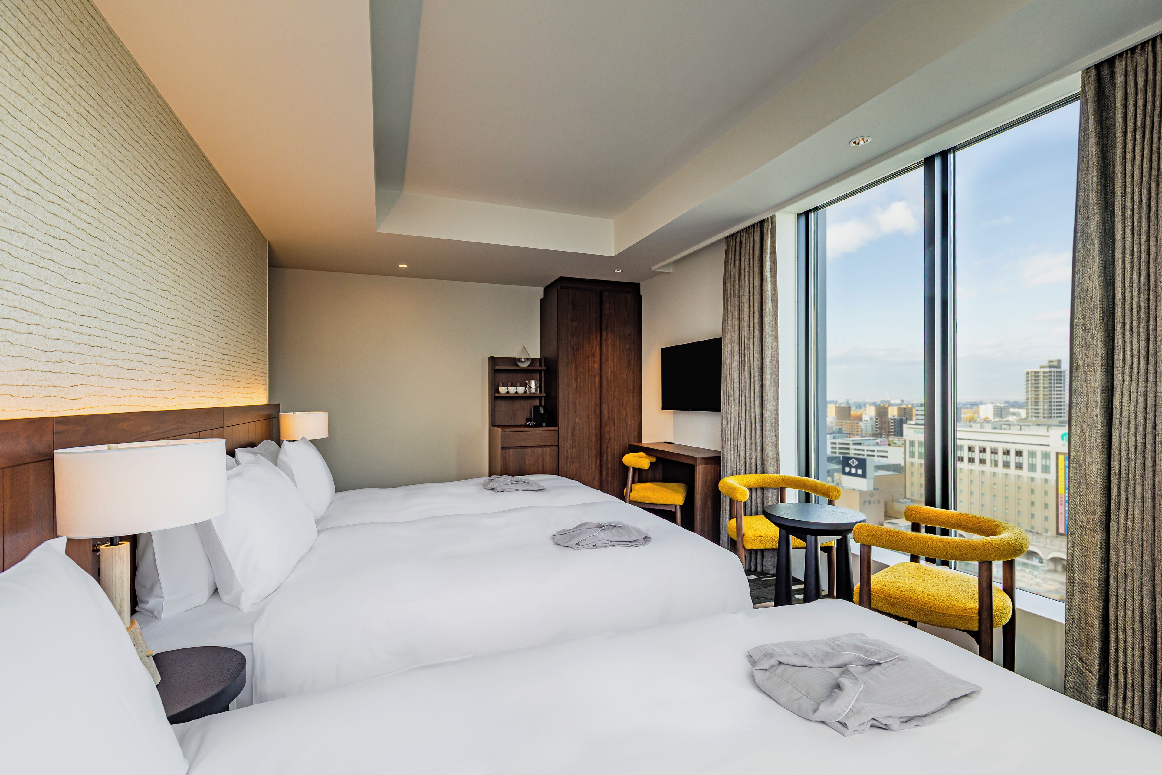 A hotel room with a large bed, a desk, two chairs, a TV, and a view of the city.