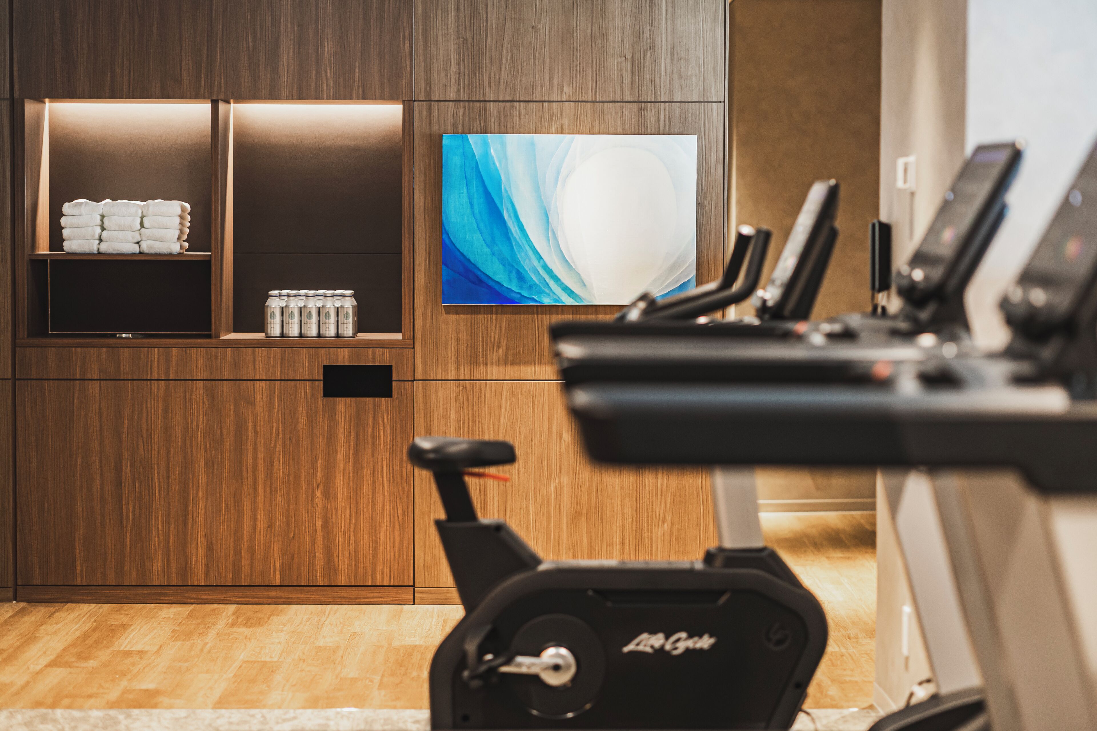 A gym with exercise equipment, a wall-mounted TV, and storage shelves with towels and beverages.