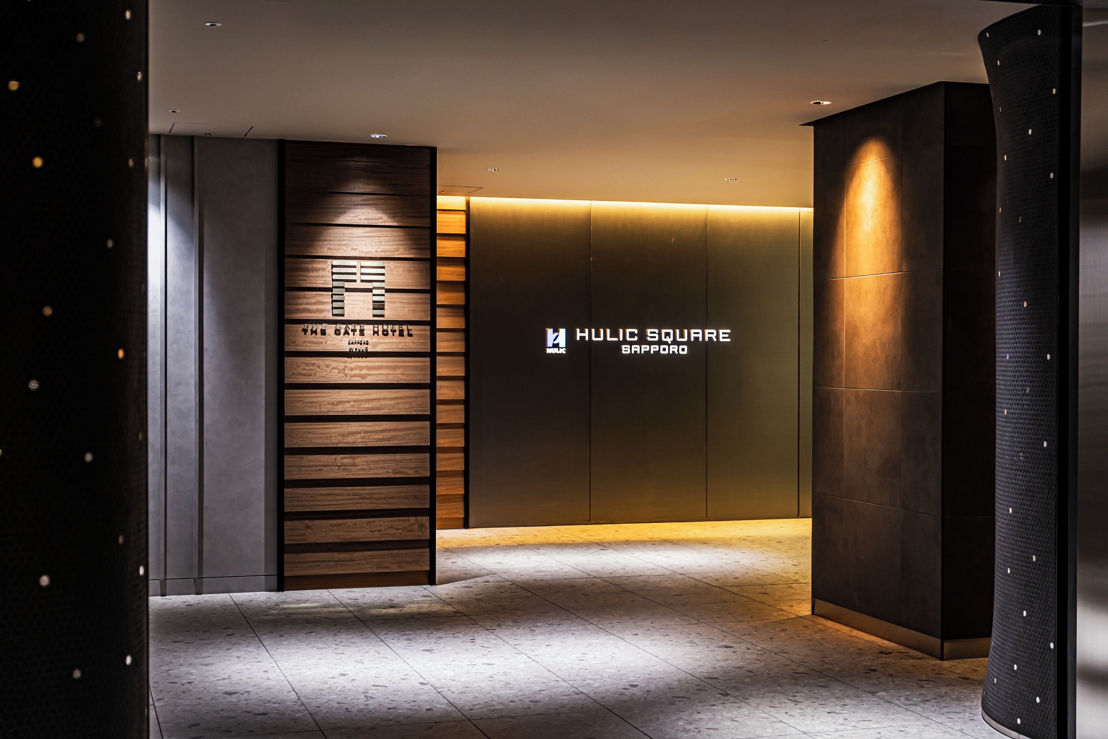 A modern hotel lobby with a wooden reception desk, a large illuminated sign reading 'HULIC SQUARE SAPPORO,' and a curved wall with a textured pattern.