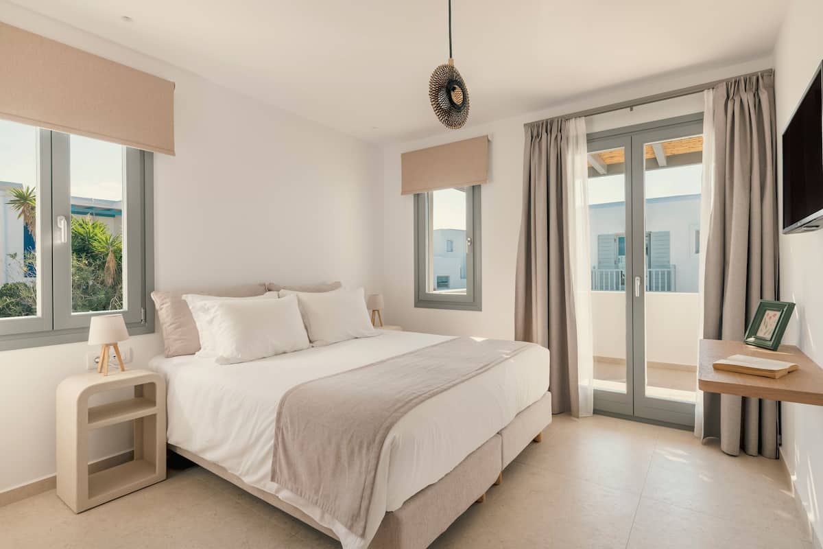 Un dormitorio moderno con una cama grande, una mesita de noche con lámpara, un televisor montado en la pared y un balcón con cortinas.