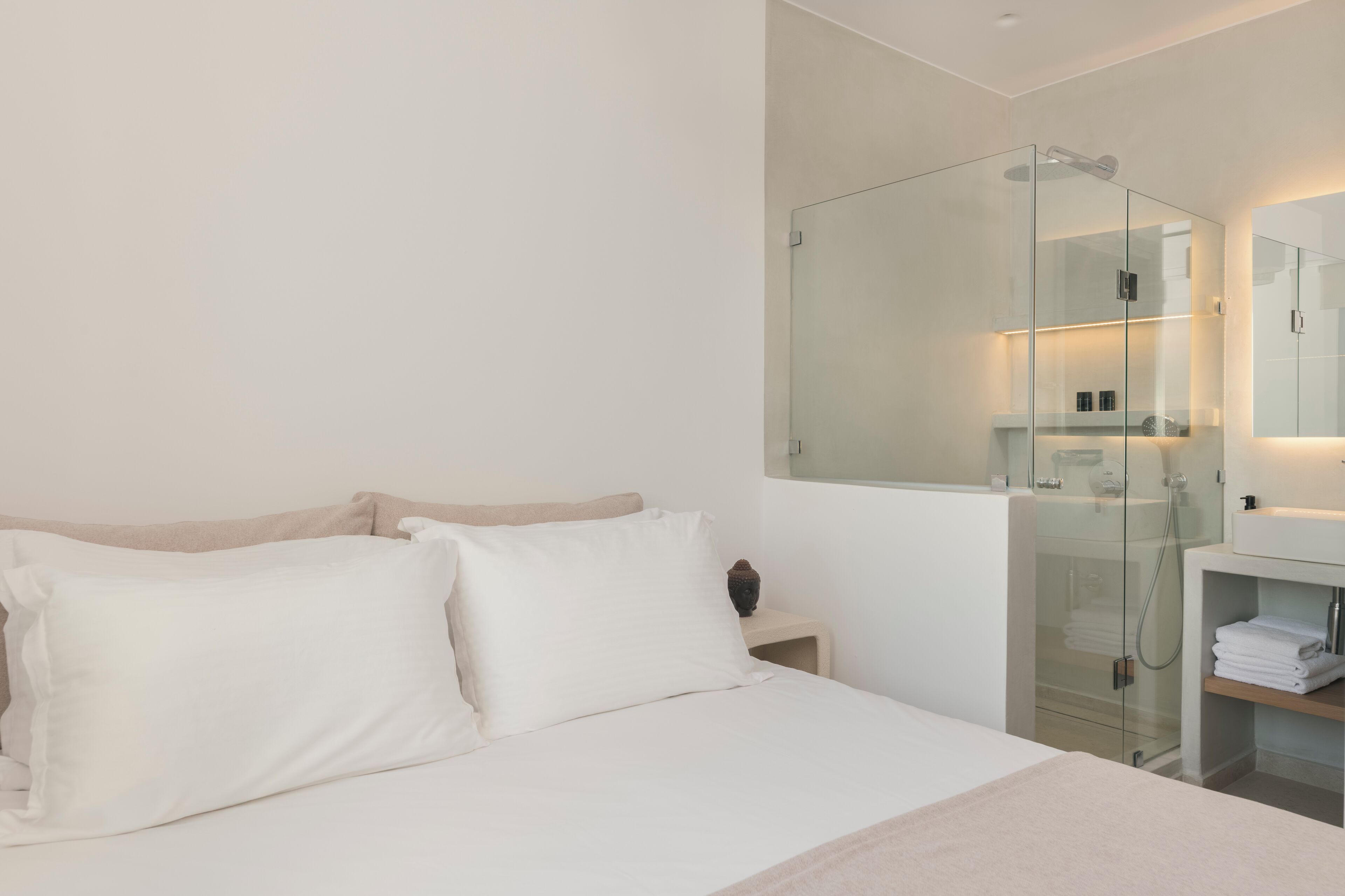 Une chambre d’hôtel moderne avec un grand lit, une douche à l’italienne en verre et une coiffeuse blanche avec un miroir.