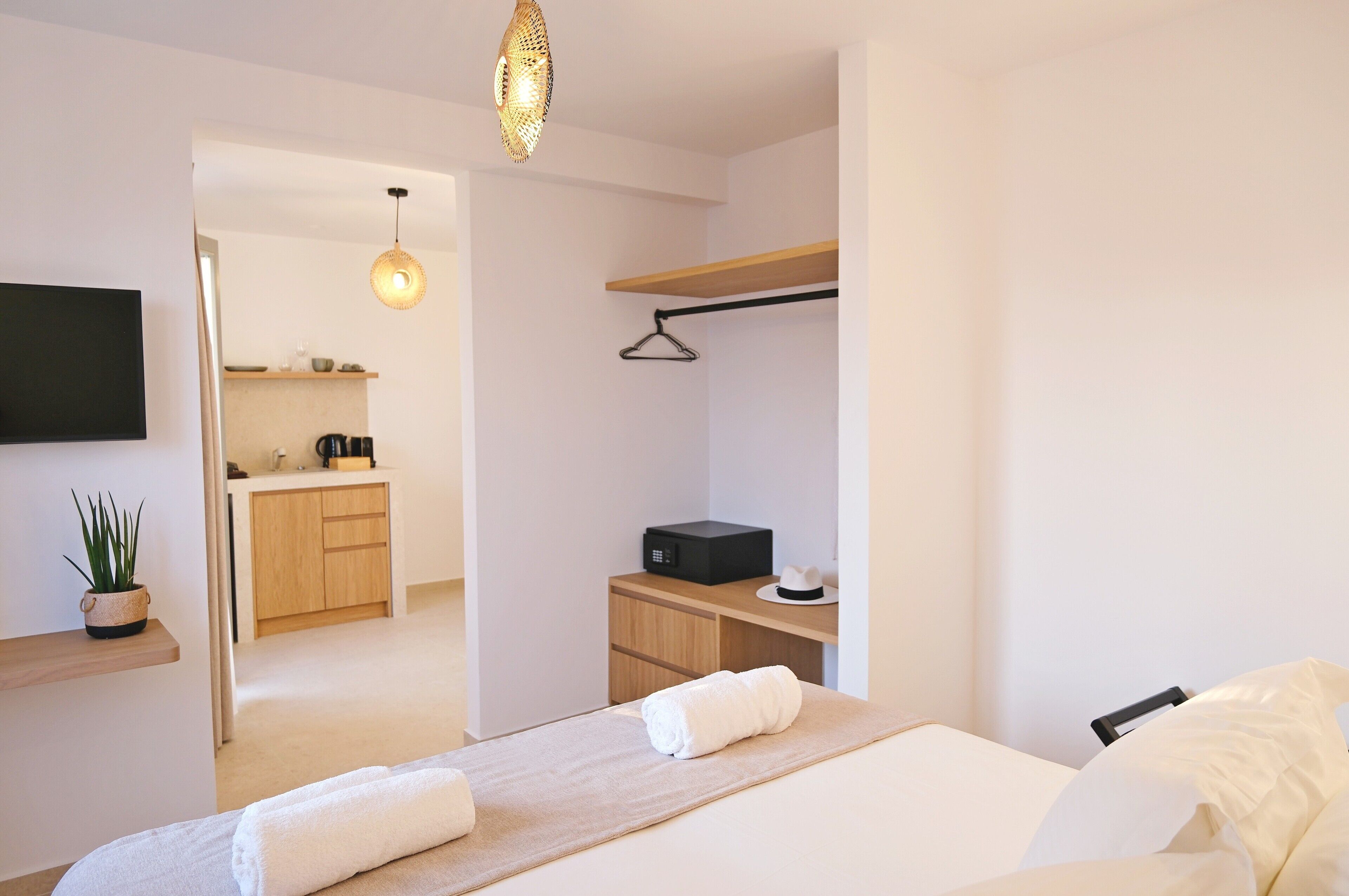 A modern hotel room with a bed, a bedside table, a hanging light fixture, a TV mounted on the wall, a small kitchen area, and a wooden shelf.