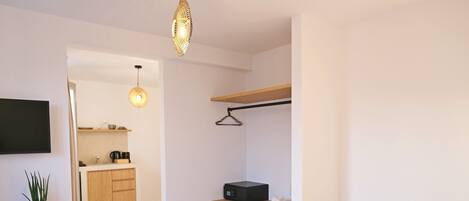 A modern hotel room with a bed, a bedside table, a hanging light fixture, a TV mounted on the wall, a small kitchen area, and a wooden shelf.