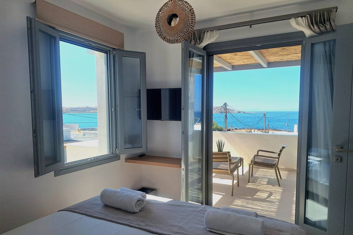 Un dormitorio con cama, dos ventanas, televisión y balcón con vista al mar.