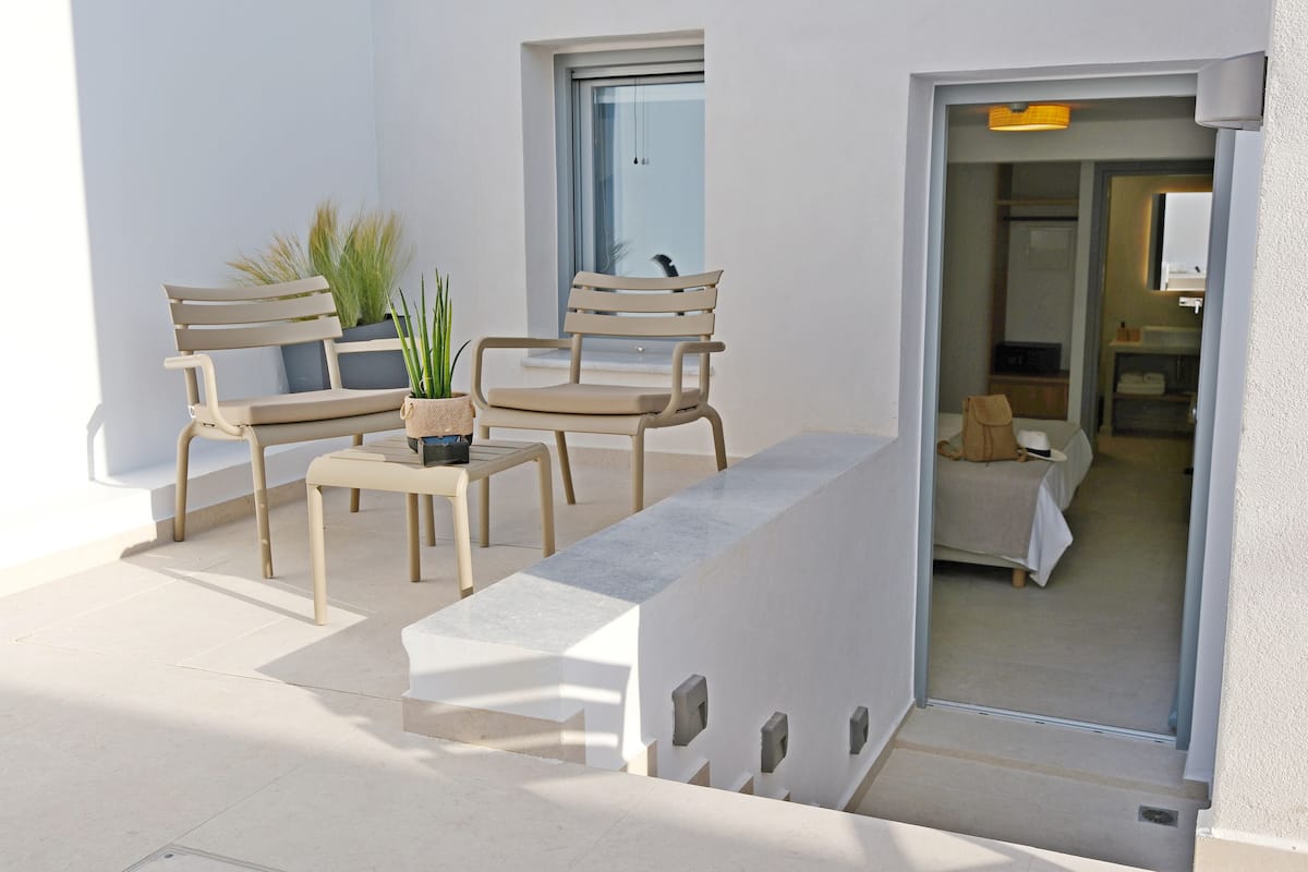 Un área de estar al aire libre y de estilo moderno, con dos sillas de madera y una mesa, una planta en maceta, y vistas a una habitación con una cama y una mesita de noche.