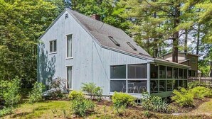 Une maison à deux étages, dotée d’une grande extension à fenêtres, et entourée de verdure.