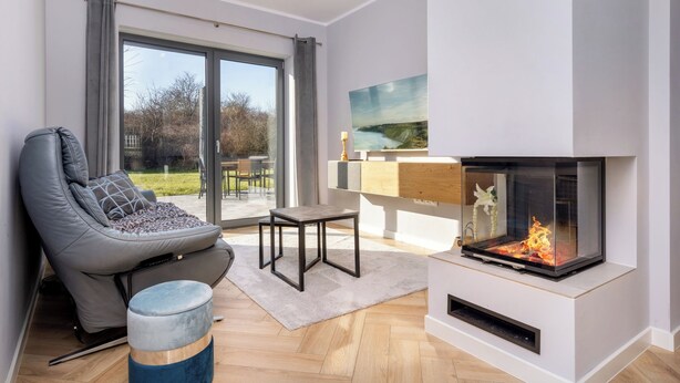 A modern living room with a fireplace, a comfortable sofa, a small table, and a view through a glass door. - Cozy vacation home Elisa, house with fireplace, sauna, terrace, garden (Dranske-Lancken)