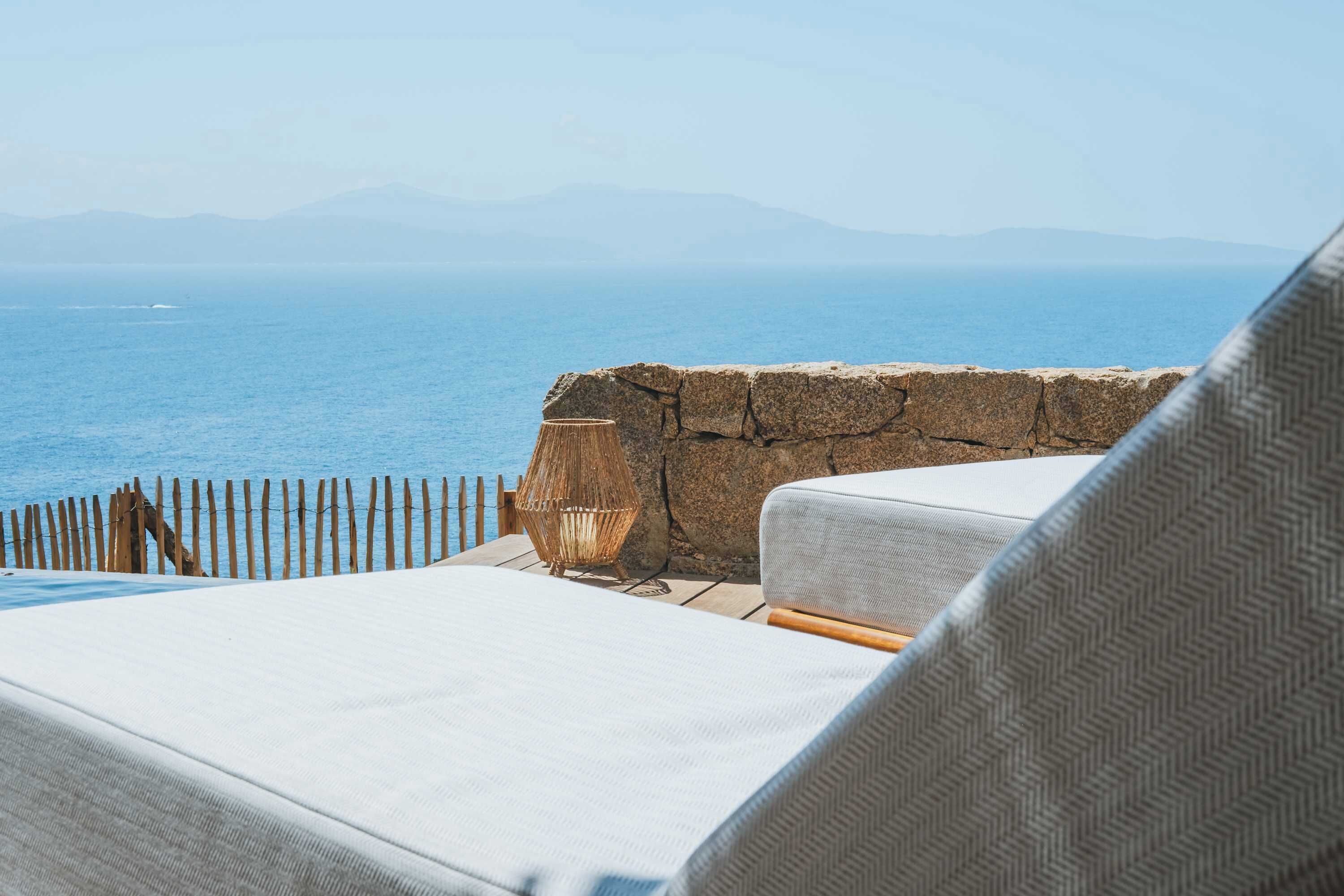 A seaside view with a stone wall, a wooden fence, and a white cushioned lounge chair.