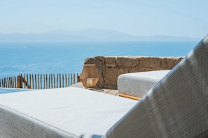 A seaside view with a stone wall, a wooden fence, and a white cushioned lounge chair.