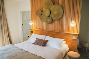 Een net opgemaakt bed met witte lakens en een bruin kussen, een houten hoofdbord met een ronde wanddecoratie, en twee wandlampen.