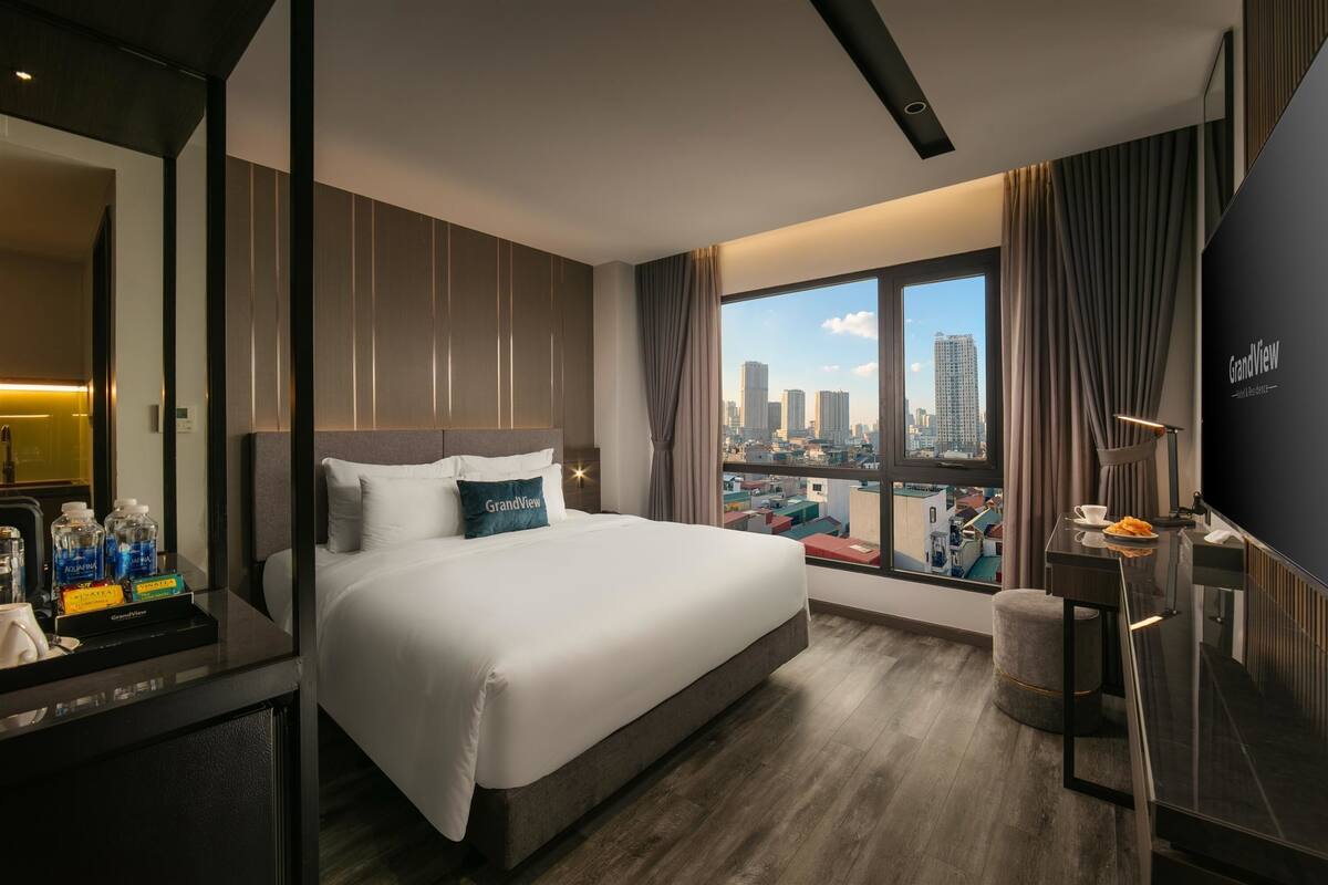 A modern hotel room with a large bed, a desk with a flat-screen TV, and a city view through the window.