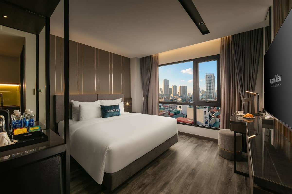 A modern hotel room with a large bed, a desk with a flat-screen TV, and a city view through the window.
