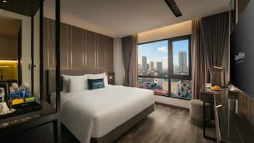 A modern hotel room with a large bed, a desk with a flat-screen TV, and a city view through the window.