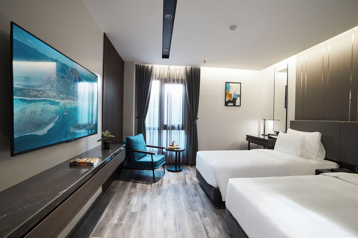Habitación de hotel con un televisor de pantalla plana grande, dos camas, un escritorio, una silla y un ventanal con cortinas.