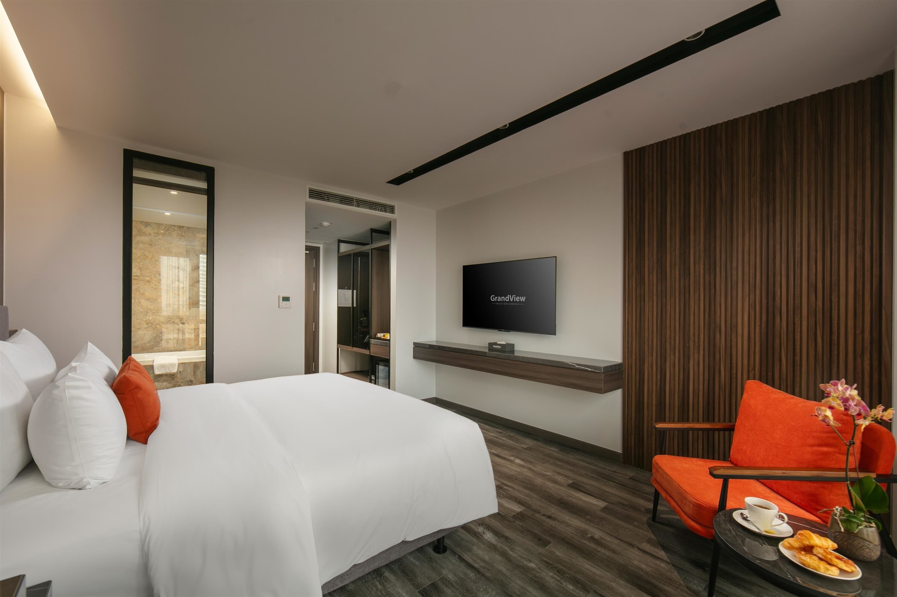 A modern hotel room with a large bed, a television mounted on the wall, a seating area with a chair and table, and a wooden panel wall.