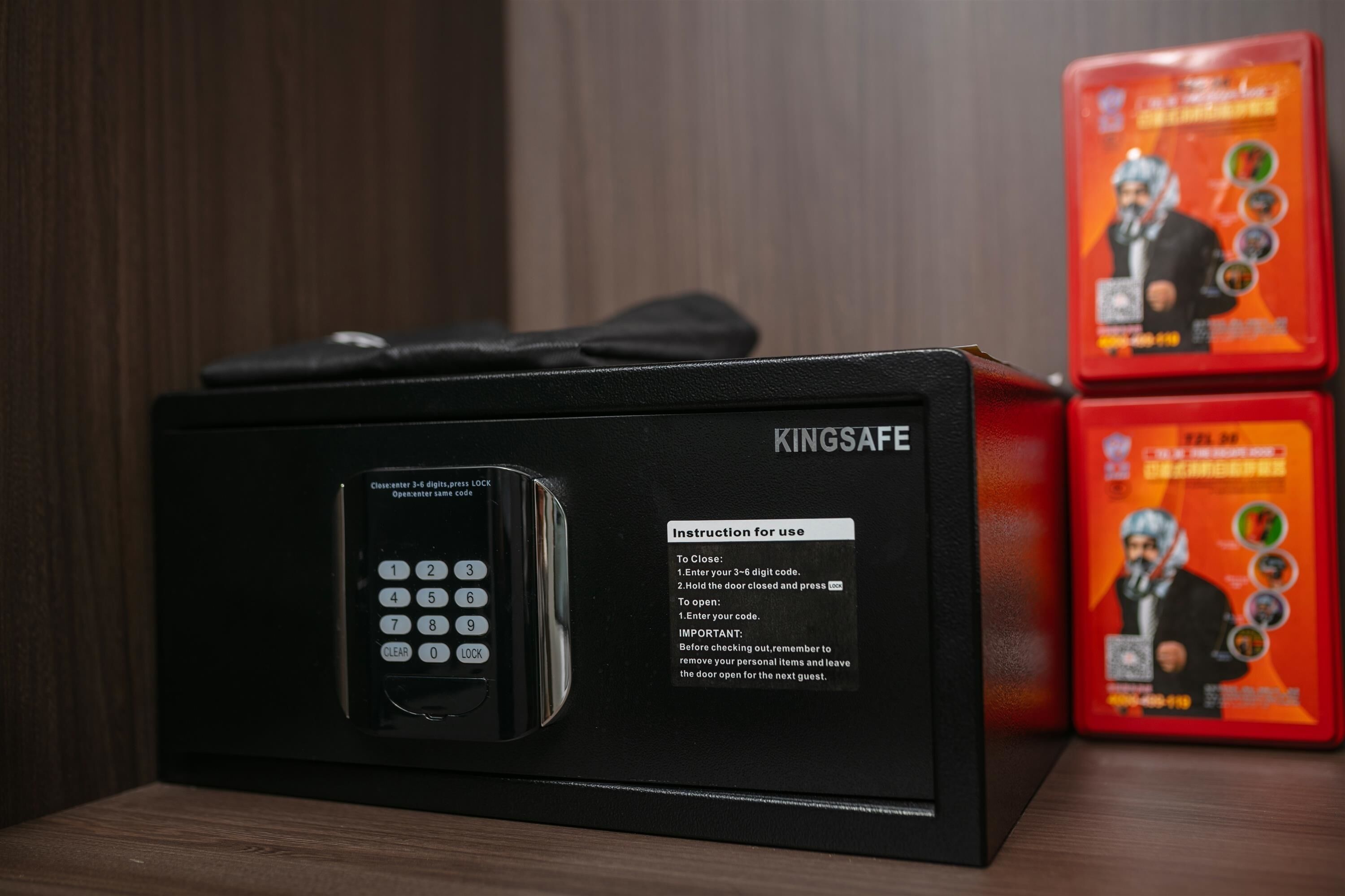 A black Kingsafe electronic lockbox with instructions for use, placed on a wooden surface with two red boxes in the background.