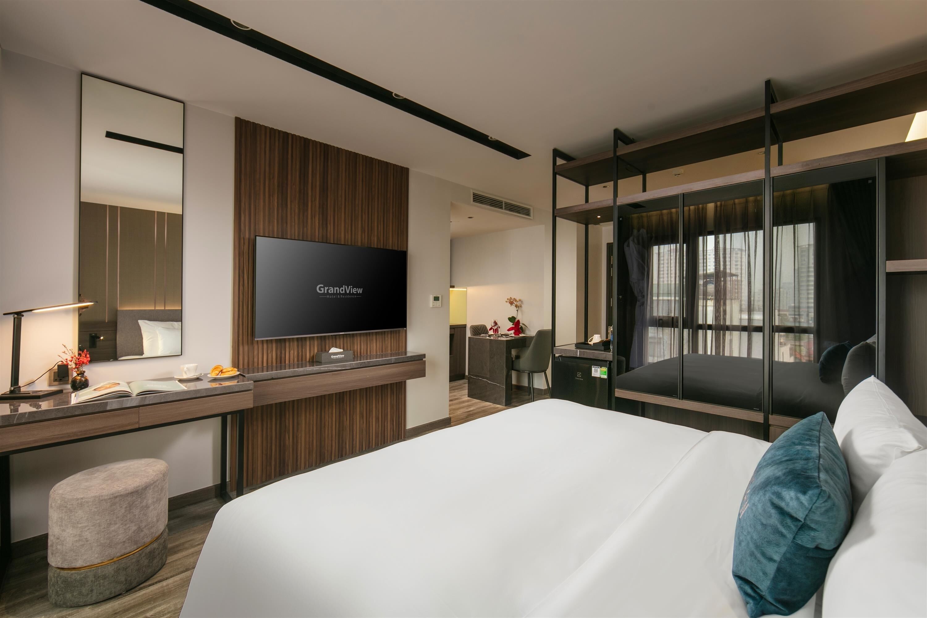 A modern hotel room with a large bed, a desk with a flat-screen TV, a seating area with a chair and a small table, and a large window with curtains.