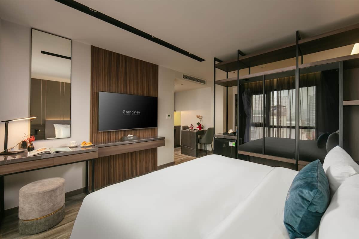 A modern hotel room with a large bed, a desk with a flat-screen TV, a seating area with a chair and a small table, and a large window with curtains.