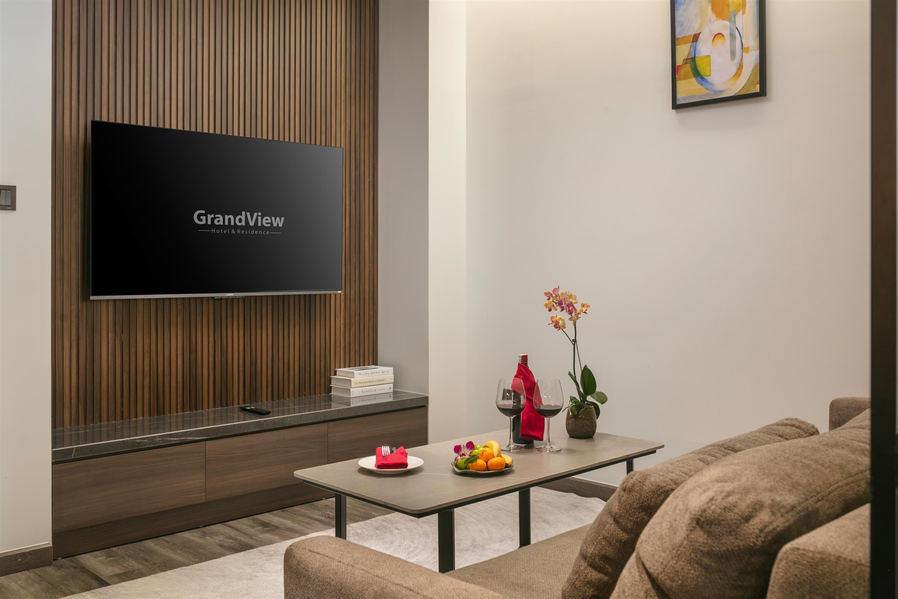 A modern living room with a flat-screen TV, wooden panel wall, a dining table with a fruit bowl, and a sofa.