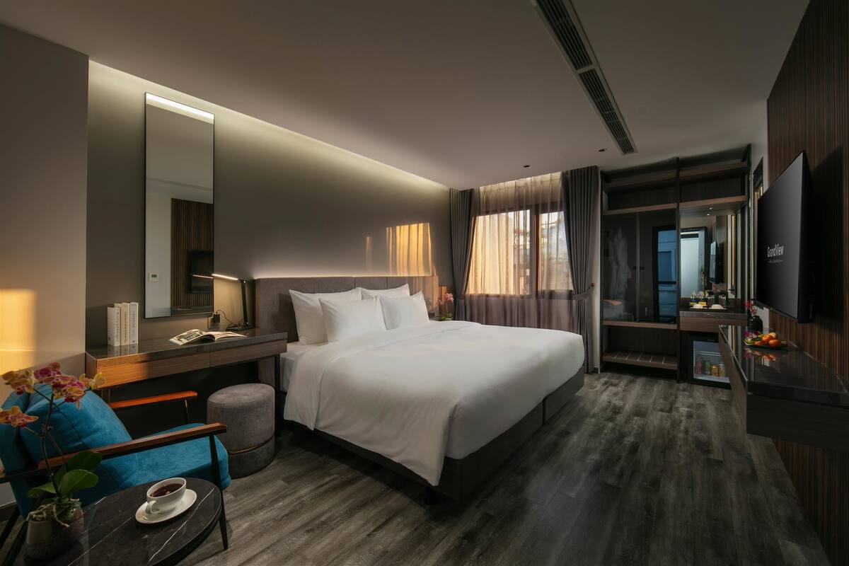 A modern hotel room with a large bed, a desk with a lamp, a chair, a small table with a cup, and a TV.