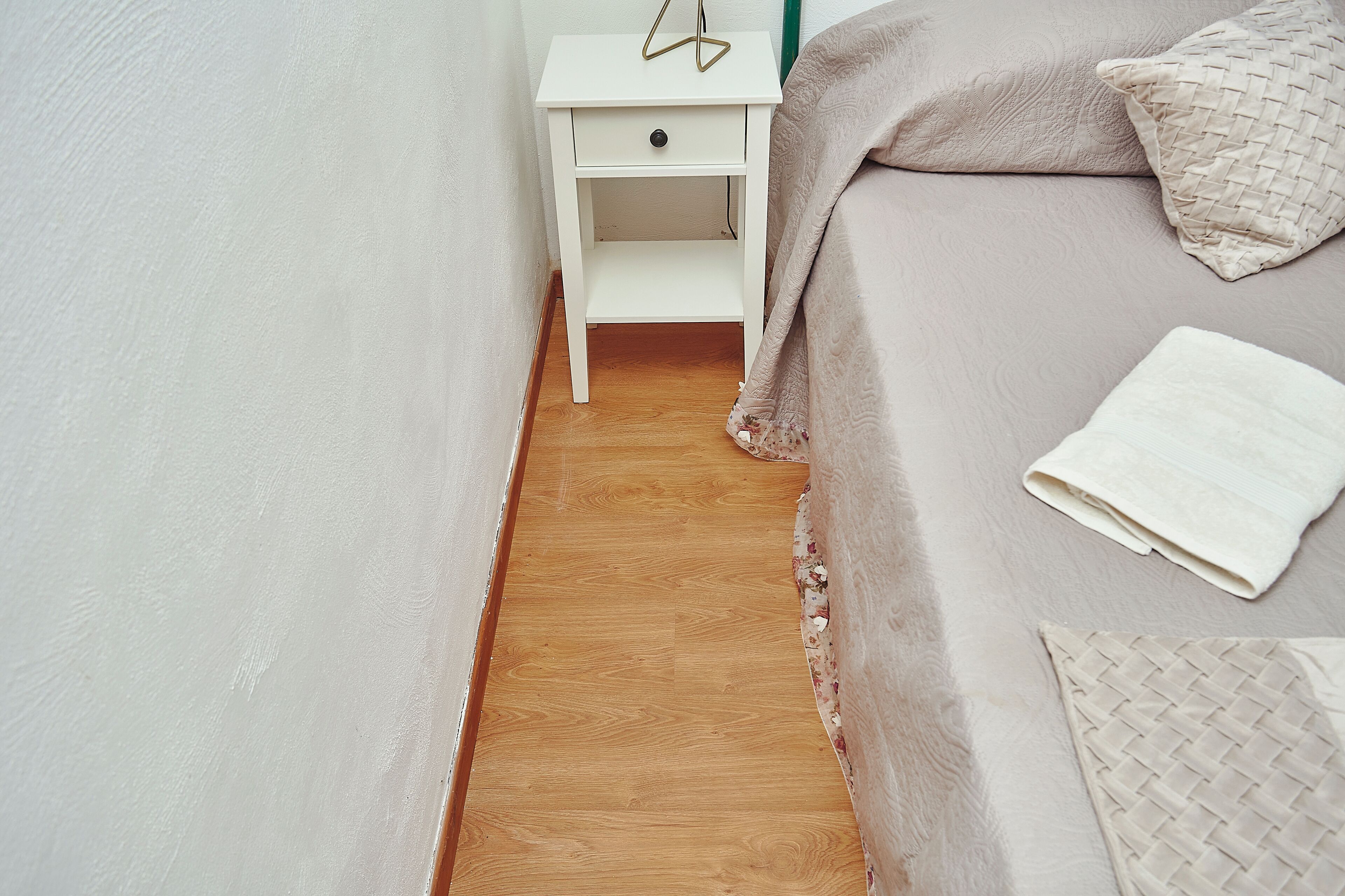 Un dormitorio con piso de madera, una mesita de noche blanca con un cajón, una cama con frazada color beige y un almohadón blanco.