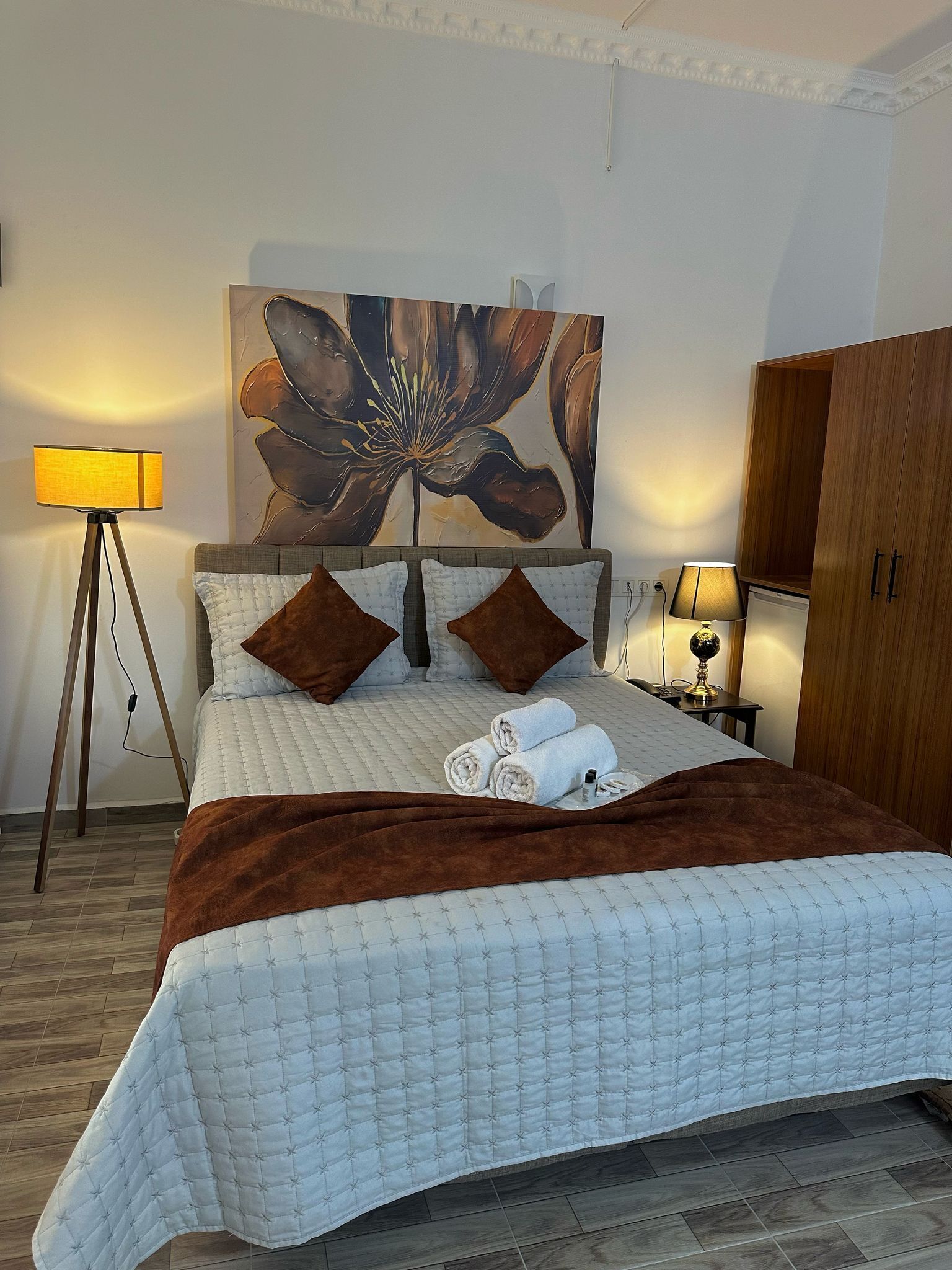 A neatly made bed with a white quilt, two brown pillows, and a brown throw blanket. There is a large floral painting on the wall above the bed, a floor lamp, and a wooden wardrobe.