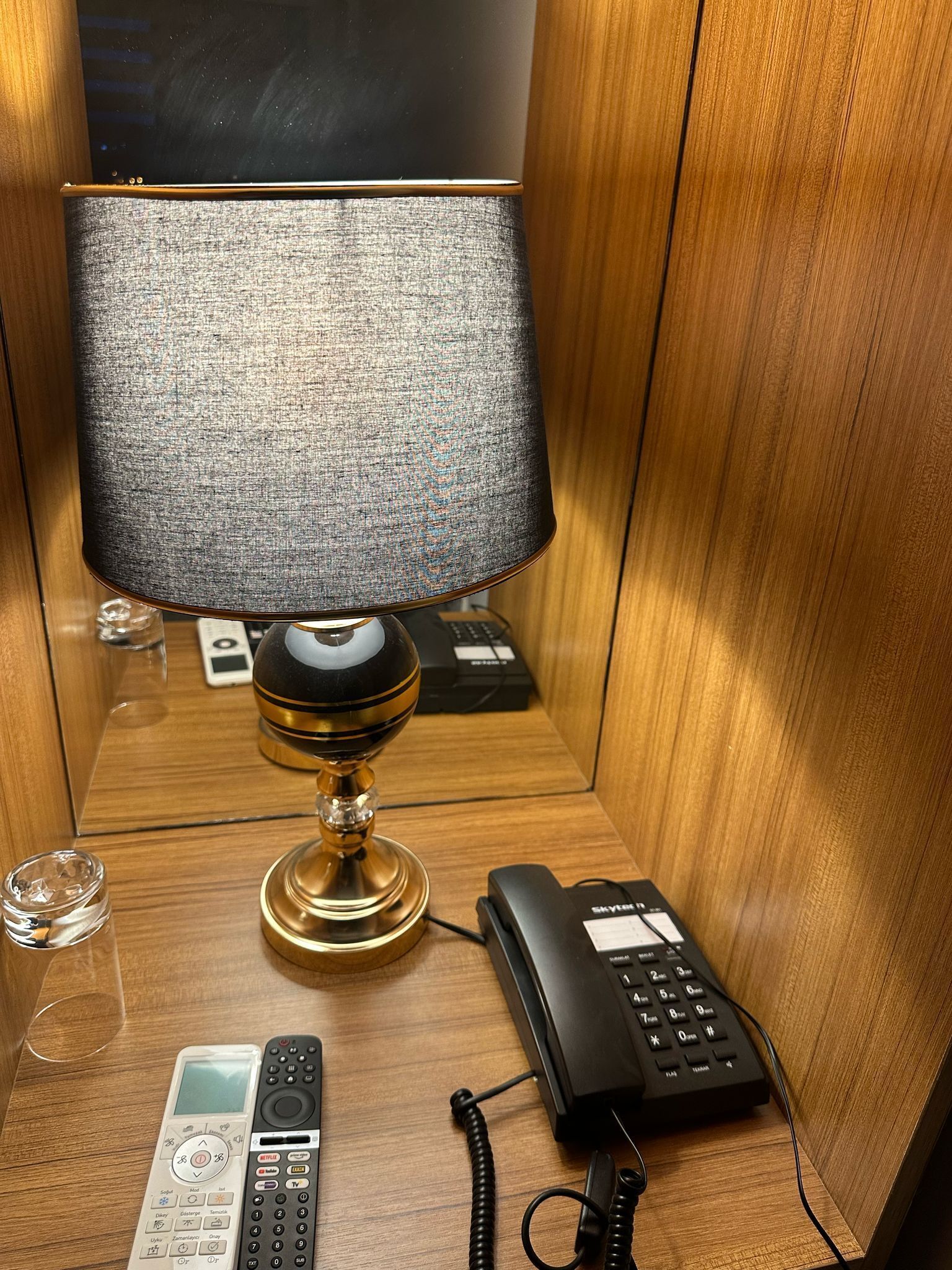 Una mesita de noche de madera con una lámpara, un teléfono y un control remoto.