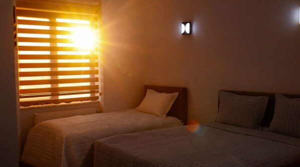 Une chambre d’hôtel avec deux lits, une fenêtre avec des stores et un luminaire fixé au mur.
