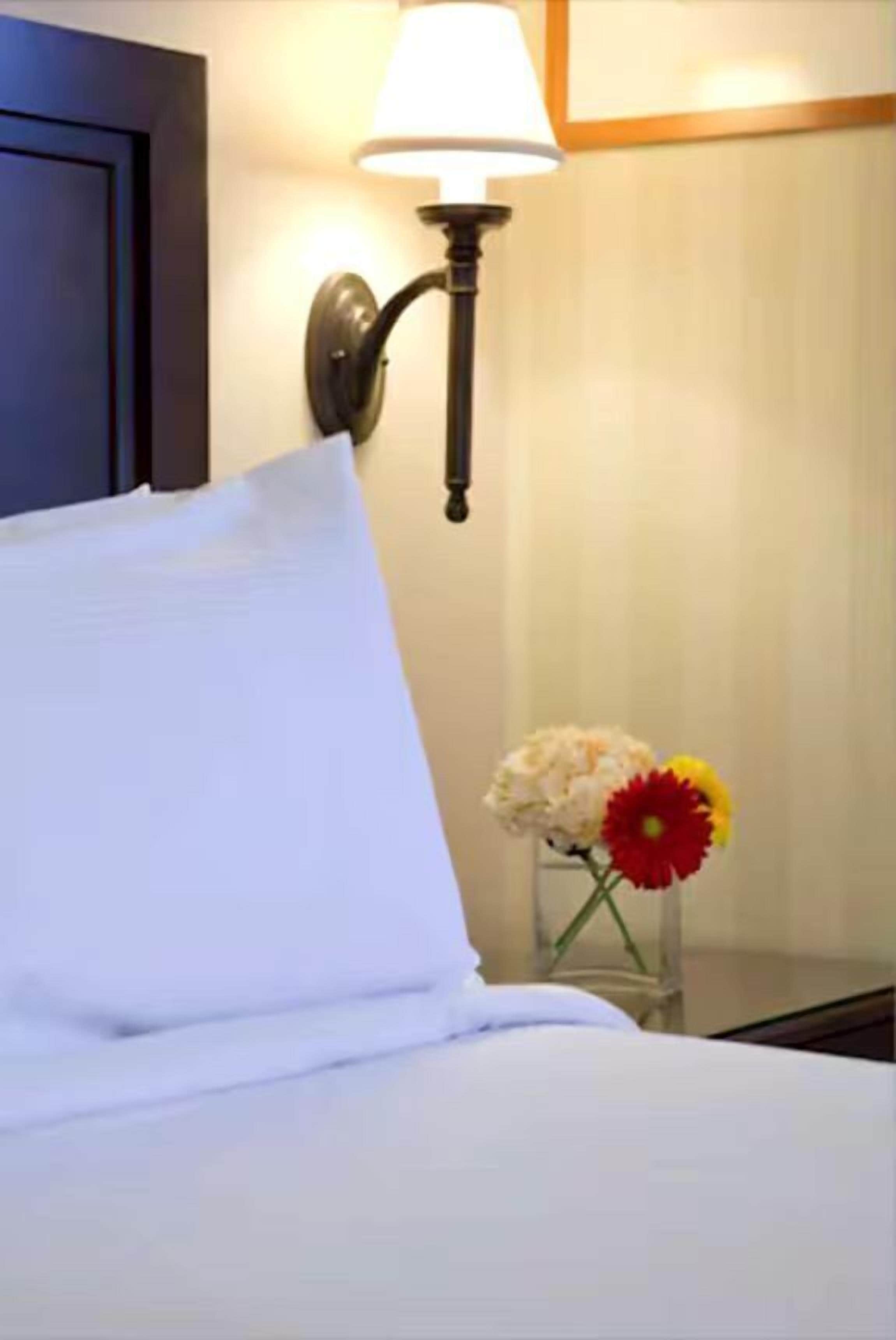 Una cama bien tendida con sábanas blancas, una lámpara de pared y un florero con flores sobre una mesita de noche.