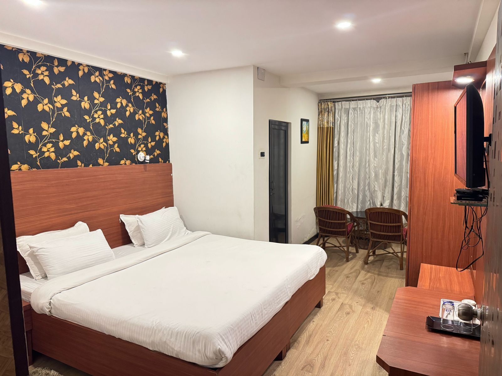 A hotel room with a large bed, a wooden headboard, a TV, a desk with a coffee maker, and a dining area with chairs and a table.