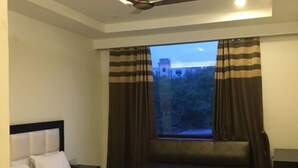 A hotel room with a bed, a window with curtains, a ceiling fan, and a view of a building outside. - Sandalwood (Meerut)