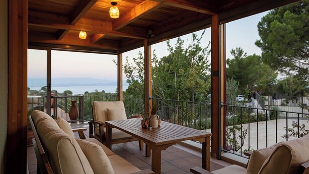 A covered outdoor seating area with wooden furniture and large windows offering a view of the surrounding landscape. - Ida Trio (Edremit)