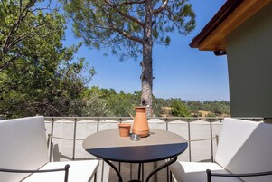 Un patio al aire libre con una mesa redonda y dos sillas blancas, una maceta de terracota y vistas a un árbol alto y un paisaje lejano.