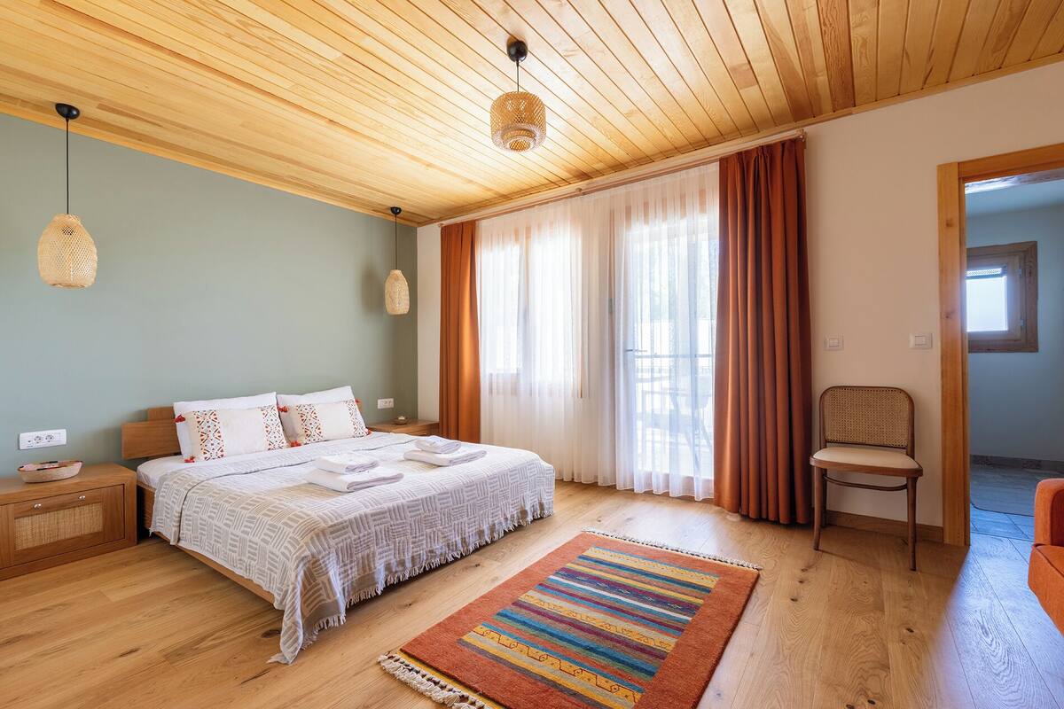 A bedroom with a wooden ceiling, a bed with white bedding, a striped rug, and a chair.