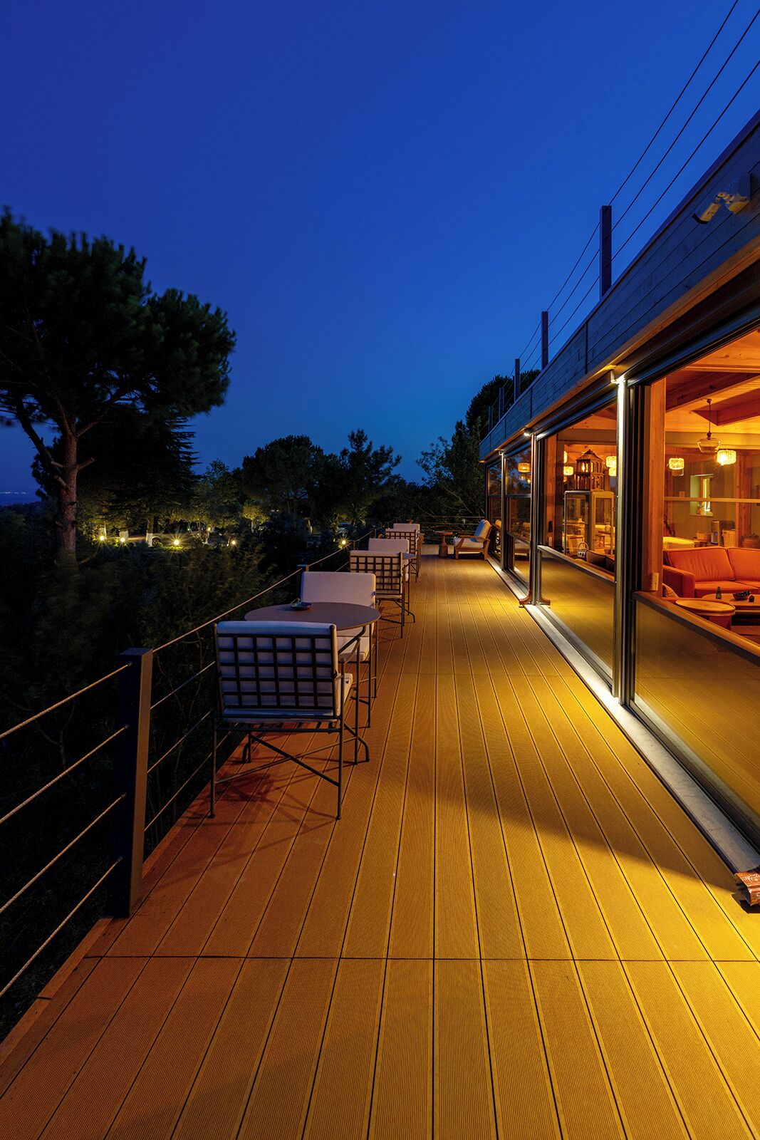 Una terraza de madera con mobiliario exterior, una habitación con paredes de vidrio y vista al cielo.