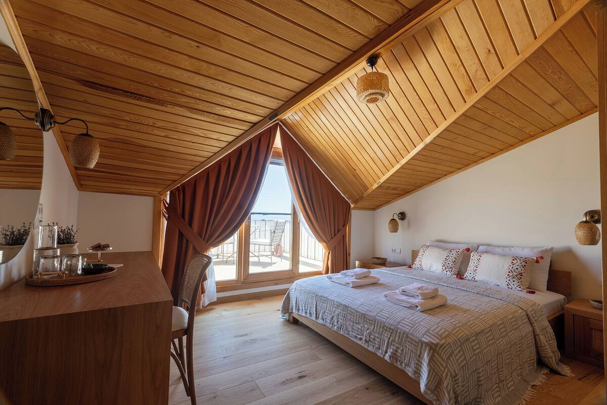 A bedroom with a sloped ceiling, wooden flooring, a bed with a quilt, a nightstand, a chair, and a window with curtains.