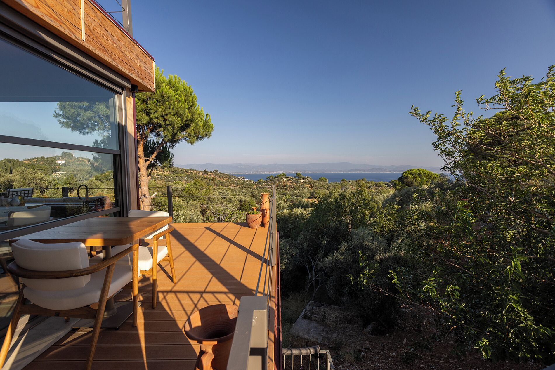 Une terrasse en bois, agréée de mobilier d’extérieur, qui offre une vue imprenable sur un paysage pittoresque, avec des arbres et une vue lointaine sur la mer.
