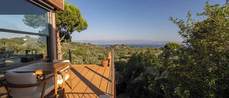 Une terrasse en bois, agréée de mobilier d’extérieur, qui offre une vue imprenable sur un paysage pittoresque, avec des arbres et une vue lointaine sur la mer.