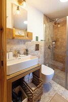 Un baño moderno con lavamanos blanco, espejo y retrete. Incluye una mampara de ducha de vidrio y un mueble de tocador de madera con una cesta.