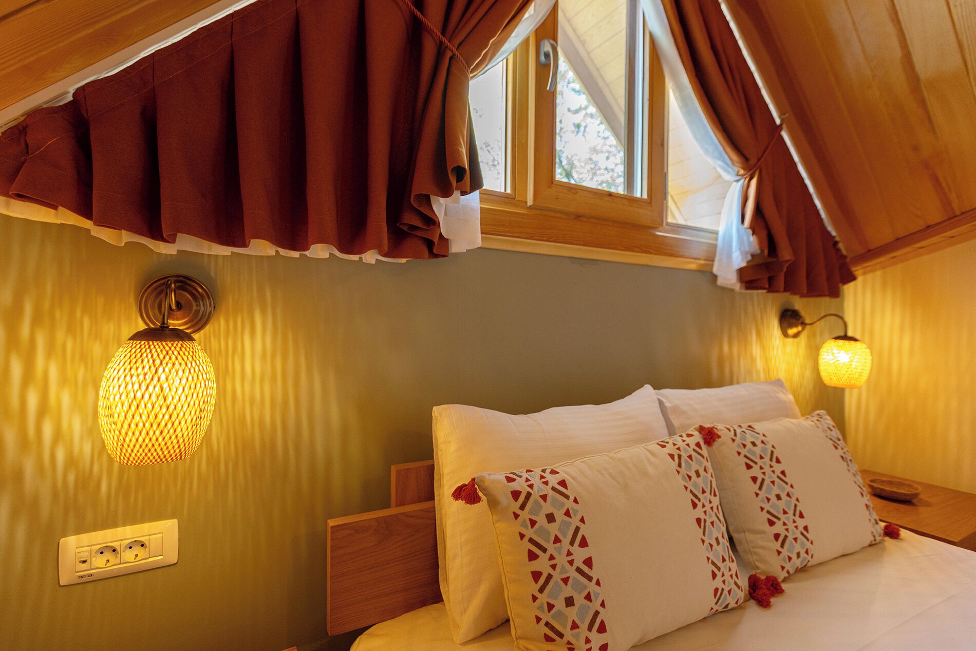 A bedroom with a wooden headboard, a bed with white linens and patterned pillows, a wall-mounted light fixture, and a window with curtains.