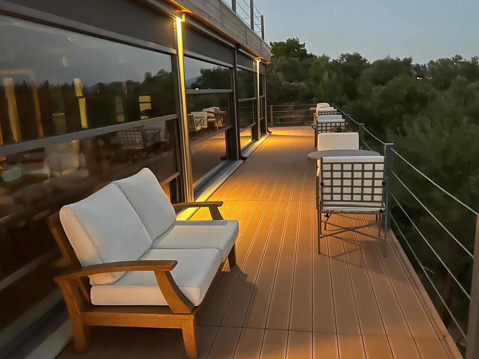 Une terrasse extérieure avec un plancher en bois et des espaces aménagés pour s’asseoir, notamment un canapé et des fauteuils, donnant sur une zone boisée.